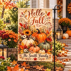 Pode incluir: Uma bandeira de jardim retangular com o texto Hello Fall e The Smith Family em letra cursiva. O design apresenta uma coleção de abóboras laranja, brancas e verdes, girassóis, bagas vermelhas e folhas de outono num fundo creme.