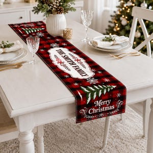 May include: A rectangular Christmas table runner featuring a red and black buffalo plaid pattern, white snowflake accents, green pine tree graphics, and the text "Merry Christmas" along with a central white label reading "THE SMITH FAMILY."