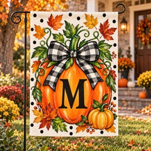 May include: A rectangular garden flag featuring a large orange pumpkin with a black and white buffalo check bow and the letter M in the center. The design includes autumn leaves, berries, and a smaller pumpkin on a white background with black dots.