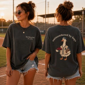 May include: Dark gray t-shirt with the text "Boy Mama Club Est. 2026" on the front. The back of the shirt features a cartoon duck wearing a baseball cap and jacket, holding a toy car and football, with the words "outnumbered by boys".