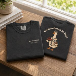 May include: Two dark gray t-shirts on a wooden surface. One shirt has "Boy Mama Club Est. 2026" printed on the front. The other shirt features a cartoon goose wearing a baseball jacket and holding a football and a toy car, with the text "outnumbered by boys".