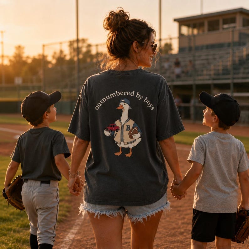 May include: A dark gray t-shirt with the text "outnumbered by boys" and a cartoon goose wearing a baseball jacket, holding a football and a red car. The image shows a woman and two children walking on a baseball field.