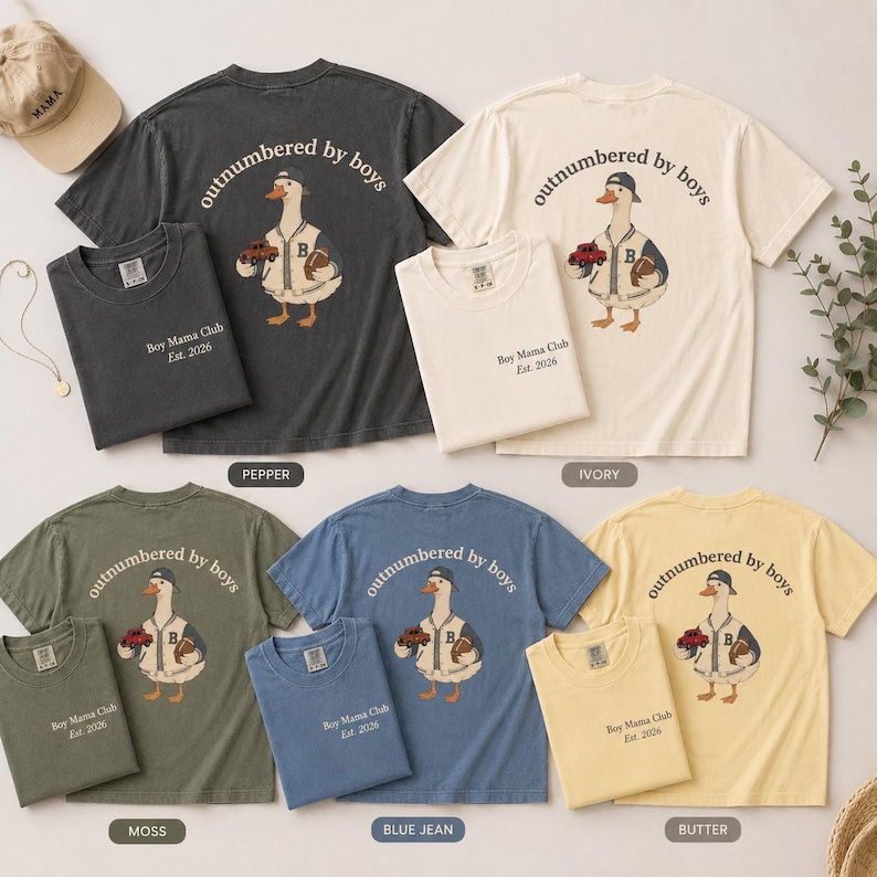 May include: A collection of t-shirts in various colors, including pepper, ivory, moss, blue jean, and butter. Each shirt features a graphic of a duck wearing a baseball cap and holding a football and a toy car. The text "outnumbered by boys" is arched above the duck, and "Boy Mama Club Est. 2026" is printed below.