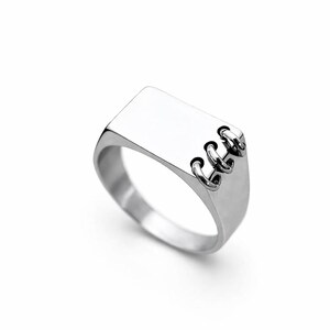 May include: A silver ring with a rectangular face and three small, connected circular accents on the side. The ring has a polished, reflective surface and a simple, modern design.