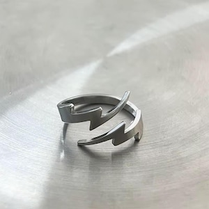 May include: A silver-colored ring with a lightning bolt design. The ring wraps around the finger with two lightning bolt shapes facing opposite directions. The ring is made of a shiny, metallic material.