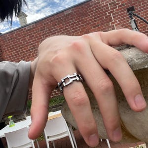 May include: A silver ring with a geometric chain link design. The ring is worn on a finger. A brick wall and cloudy sky are in the background. The ring's design features interconnected rectangular shapes.
