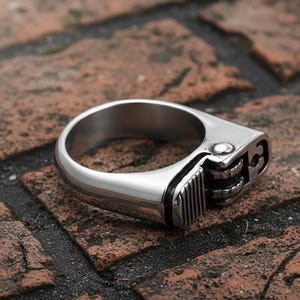 May include: A silver-colored ring designed to resemble a lighter. The ring features a detailed replica of a lighter's components, including the flint wheel, fuel adjustment, and the body of the lighter. The ring is set against a textured brick surface.