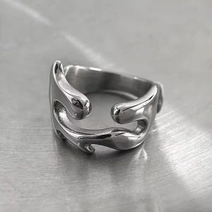 Peut inclure: Une bague argentée au design unique et fluide. La bague présente une forme incurvée et ouverte avec des surfaces lisses et polies. Le design a une esthétique fluide et organique, avec des bords arrondis et un look moderne. La bague est probablement en acier inoxydable ou en métal similaire.