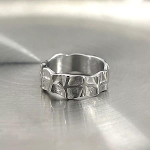 May include: A silver ring with a textured, geometric design. The ring's surface has a pattern of irregular, interconnected shapes, creating a unique and modern aesthetic. The ring is made of a shiny, metallic material.