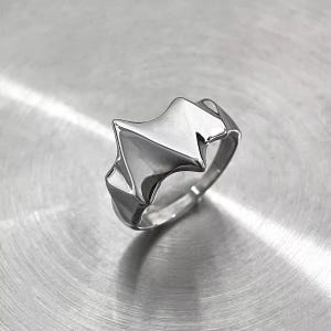 May include: A silver-colored ring with a geometric design. The ring features a central, angular shape with pointed edges, and a smooth, polished finish. The band is a simple, rounded design. The ring is displayed on a metallic surface.