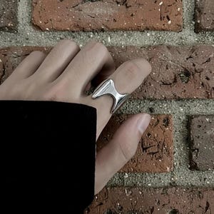 Könnte beinhalten: Ein silberfarbener Ring mit einem einzigartigen, kantigen Design. Der Ring hat ein zentrales, spitzes Element, das von zwei gebogenen, flügelartigen Formen flankiert wird. Der Ring wird an einem Finger getragen, vor einem Hintergrund aus Backsteinmauerwerk.