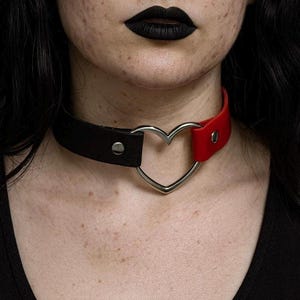 May include: A black and red choker necklace with a silver heart-shaped center. The choker is made of leather and has a buckle closure. The red section is on the right side, and the black section is on the left.