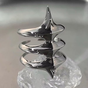 May include: A silver-colored ring with a unique, sculptural design. The ring features a spiral shape with sharp, pointed elements. The polished metal reflects light, highlighting the ring's intricate details and modern aesthetic. The ring is displayed on a clear, textured surface.