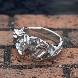 Puede incluir: Un anillo plateado con un diseño orgánico y texturizado. El anillo tiene una superficie irregular con formas abstractas y fluidas. El anillo está sobre un fondo de ladrillos rojos.