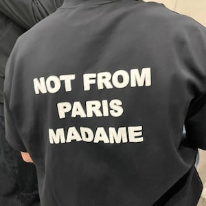May include: Black crew neck sweatshirt with the white text "NOT FROM PARIS MADAME" across the back. The sweatshirt is a solid black color. The person wearing the sweatshirt is facing away from the camera.