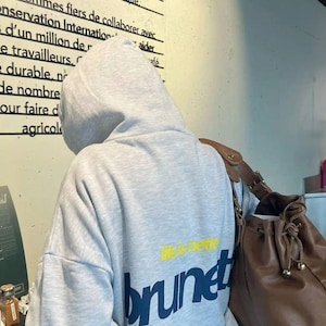 Puede incluir: Una sudadera con capucha gris claro con la palabra "brunet" en azul marino y "life is better" en amarillo. Un bolso de cuero marr&oacute;n es visible a la derecha. El fondo incluye texto en franc&eacute;s.