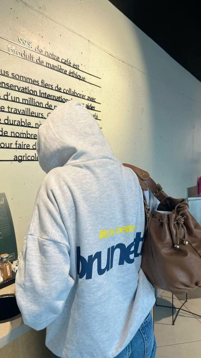 Puede incluir: Una sudadera con capucha gris claro con la palabra "brunet" en azul marino y "life is better" en amarillo. Un bolso de cuero marr&oacute;n es visible a la derecha. El fondo incluye texto en franc&eacute;s.
