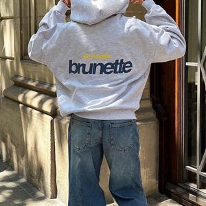 Puede incluir: Una sudadera con capucha gris claro con el texto "life is better brunette" en azul marino y amarillo. La sudadera se combina con unos vaqueros azules. La imagen est&aacute; tomada desde atr&aacute;s, mostrando el dise&ntilde;o de la sudadera.
