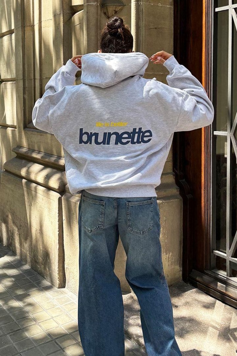 Puede incluir: Una sudadera con capucha gris claro con el texto "life is better brunette" en azul marino y amarillo. La sudadera se combina con unos vaqueros azules. La imagen est&aacute; tomada desde atr&aacute;s, mostrando el dise&ntilde;o de la sudadera.
