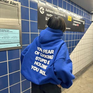 Puede incluir: Una sudadera con capucha azul real con el texto "YOU FEAR OF LOOKING STUPID IS HOLDING YOU BACK" en blanco. La persona está de pie en una estación de metro con paredes de azulejos azules y señalización.