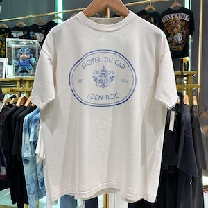 以下が含まれることがあります： 胸元に青い楕円形のグラフィックがプリントされたクリーム色の半袖Tシャツ。グラフィックには「HOTEL DU CAP」「EDEN-ROC」「EST 1870」の文字と中央に紋章のイラストが描かれています。クルーネックで標準的なフィット感のTシャツです。