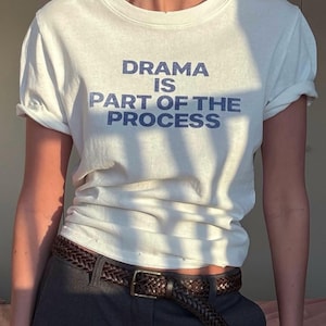 May include: A white short-sleeve t-shirt with a crew neckline featuring the dark blue text "DRAMA IS PART OF THE PROCESS" printed on the chest. The shirt is paired with dark gray trousers and a brown woven leather belt with a gold-tone metal buckle.