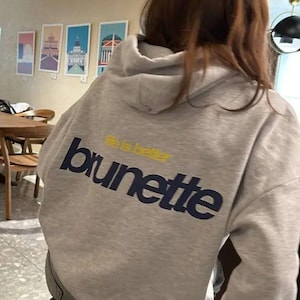 Puede incluir: Sudadera con capucha gris claro con la palabra "brunette" en letras may&uacute;sculas azul marino y las palabras "life is better" en amarillo encima. La sudadera tiene capucha y mangas largas.