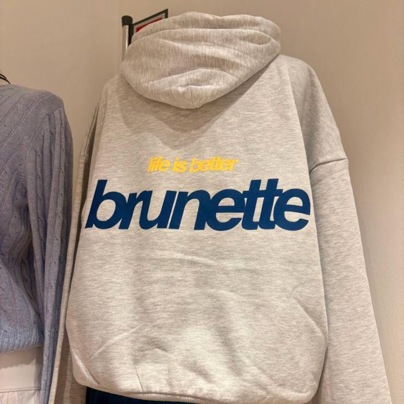 Puede incluir: Una sudadera con capucha gris claro con la palabra "brunette" en letras azules. Encima de la palabra "brunette" est&aacute; la frase "life is better" en amarillo. La sudadera tiene capucha y mangas largas.