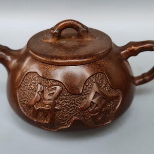 May include: A brown, antique-style teapot with a lid and a bamboo-style handle. The teapot has carved Chinese characters on the side. The spout and handle are curved. The teapot is made of a dark brown material.