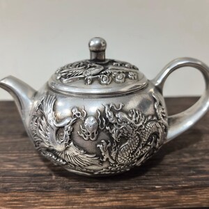 Könnte beinhalten: Eine silberne Teekanne mit aufwendigen Reliefs von Drachen und Phönixen. Die Teekanne hat einen gebogenen Ausguss, einen abgerundeten Griff und einen Deckel mit einem kleinen Knopf. Das detaillierte Design deutet auf eine asiatisch inspirierte Dekoration hin.