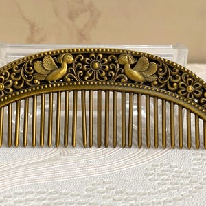Puede incluir: Un peine para el cabello de estilo antiguo con acabado bronce. El peine presenta un diseño arqueado con intrincados motivos florales y de aves. Los dientes del peine están espaciados uniformemente, lo que sugiere un accesorio para el cabello vintage o decorativo.