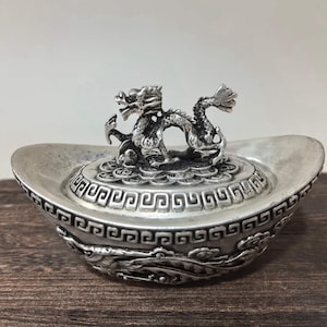 May include: A silver-colored ingot-shaped decorative item with an ornate dragon figurine on top. The dragon is detailed with scales and a flowing tail. The ingot has decorative patterns around the edges.