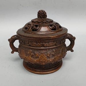 May include: A bronze-colored incense burner with a lid and two handles. The lid is intricately carved with openwork designs and a decorative knob. The body of the burner features detailed relief carvings. The handles are shaped like dragon heads.