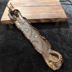 Puede incluir: Un porta incienso de color bronce en forma de dragón. La cabeza y la cola del dragón están en los extremos opuestos, con un cuerpo detallado con escamas y motivos de nubes. Descansa sobre un pequeño soporte de madera.