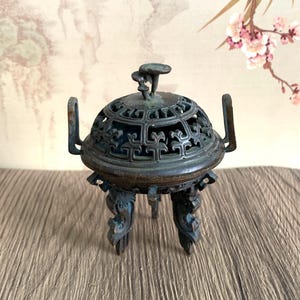 May include: A bronze incense burner with a domed, pierced lid and ornate handles. The burner rests on three decorative legs. The background features a painting of a landscape and cherry blossoms.