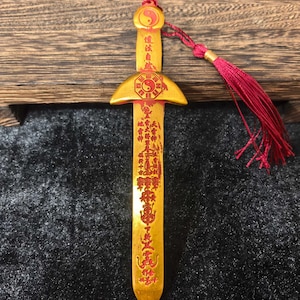 Puede incluir: Una espada decorativa dorada con caracteres y símbolos chinos rojos. La espada presenta una borla roja y un símbolo de yin-yang. La hoja está inscrita con texto vertical. La espada se exhibe sobre una superficie oscura.