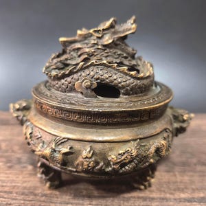 Puede incluir: Un quemador de incienso de metal en tono bronce con una tapa rematada por una escultura de un dragón enroscado. El cuerpo redondo del recipiente está decorado con relieves de dragones y patrones geométricos, apoyado sobre tres patas.