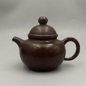 May include: A brown ceramic teapot with a rounded body, a curved handle, and a small spout. The lid has a small, round knob. The teapot is a solid, dark brown colour and appears to be made of glazed ceramic.