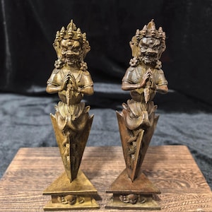 Puede incluir: Dos estatuas de color bronce de una deidad con corona y expresión feroz. Las figuras sostienen una cuchilla y están montadas sobre una base triangular con detalles de calaveras. Las estatuas se exhiben sobre una superficie de madera.