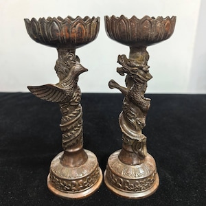 Könnte beinhalten: Zwei antike Kerzenhalter aus Bronze. Einer zeigt einen Phönix, der andere einen Drachen. Beide haben verzierte Sockel und lotusförmige Kerzenschalen. Die Kerzenhalter sind dunkelbronzen.