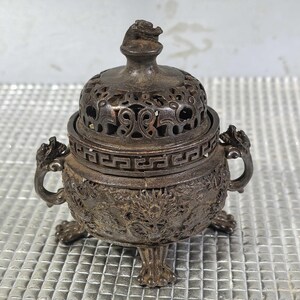 May include: A detailed, antique metal incense burner with a lid. The burner is dark silver with intricate dragon and floral patterns. It has a small dragon figure on top of the lid and dragon-shaped handles. The burner stands on three clawed feet.