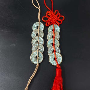 May include: Two strings of antique Chinese coins. One string is tied with a tan cord, the other with a red cord and tassel. The coins have a square hole in the center and are strung together.