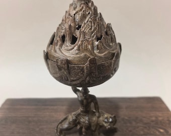 May include: A bronze-colored incense burner with a mountain-shaped lid featuring cut-out designs. The burner is supported by a figure riding a lion. The incense burner is placed on a wooden surface.
