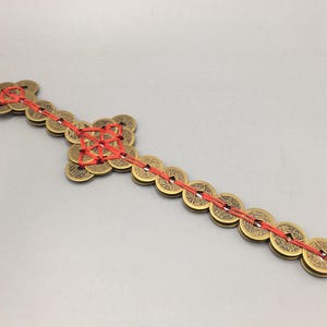 May include: A collection of bronze-colored Chinese coins strung together with red cord. The coins are round with square holes in the center. The arrangement forms a decorative pattern, likely for good luck or Feng Shui.