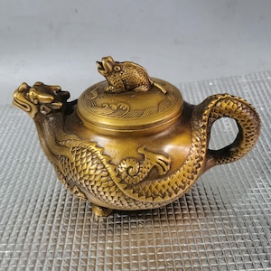 Puede incluir: Una tetera de color bronce con intrincados detalles de dragón y rana. La tetera presenta una boquilla con cabeza de dragón, un cuerpo de escamas de dragón y una figura de rana en la tapa. El asa también está diseñada con escamas de dragón.