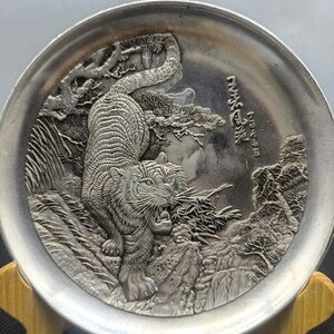 Puede incluir: Un plato decorativo plateado con un relieve detallado de un tigre en un entorno natural. El tigre se representa con la boca abierta, rodeado de árboles y rocas. Se ven caracteres japoneses en el plato.