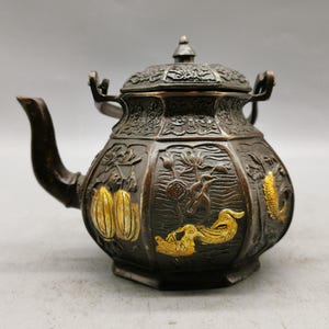 Puede incluir: Una tetera de color bronce con intrincados diseños florales y de patos, acentuada con dorado. La tetera presenta un pico curvo, una tapa decorativa y dos asas. El diseño general sugiere una estética vintage o antigua.