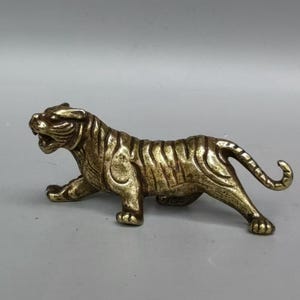 Könnte beinhalten: Eine bronzefarbene Tigerfigur in Gehposition. Die Skulptur hat eine detaillierte Fellstruktur und einen offenen Mund, der seine Zähne zeigt. Der Tiger ist auf allen Vieren, mit eingerolltem Schwanz.