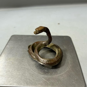 May include: A bronze-coloured snake figurine coiled on a silver-coloured surface. The snake's head is raised, and its body is textured with scales. The figurine is a decorative item.
