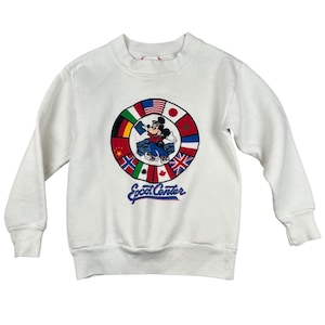 Può includere: Felpa bianca con grafica circolare di Topolino in un'auto circondata da bandiere di diversi paesi. Il testo "Epcot Center" è stampato in blu. La felpa ha girocollo e maniche lunghe.